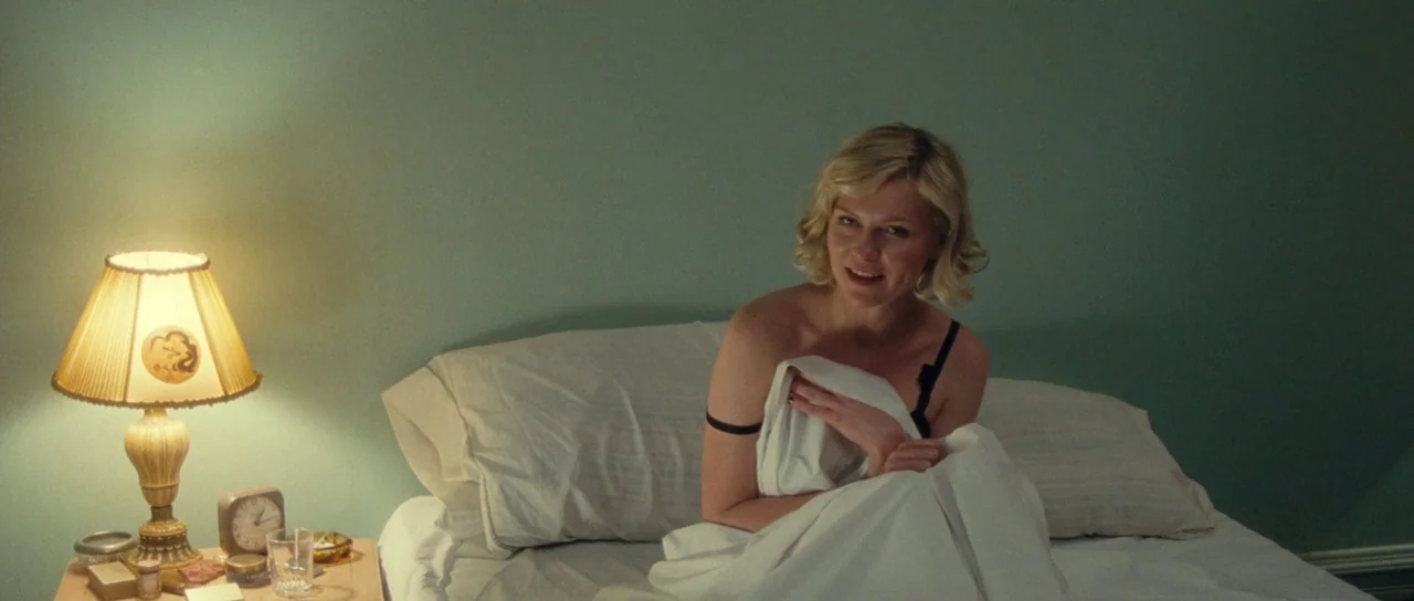 On The Road (2012) movie still: medium — A bedroom with muted green walls and a bedside table with a lamp. A woman sits in bed covered by a white…