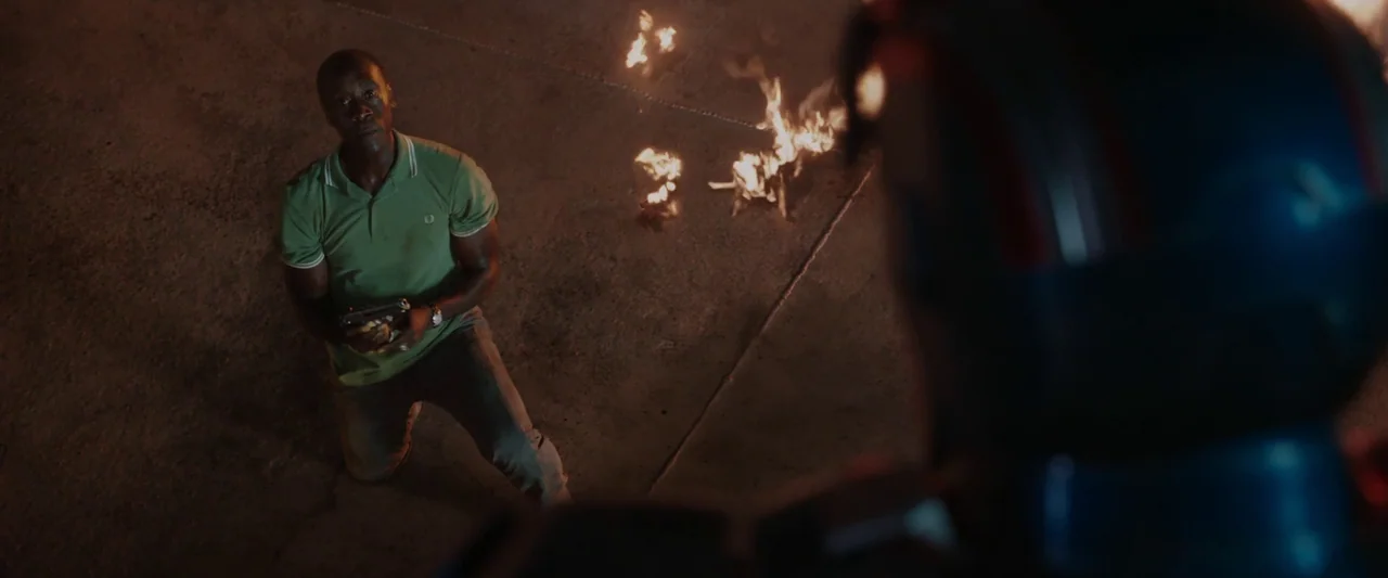 Iron Man 3 (2013) movie still: medium — A man kneels on an asphalt surface at night near flickering flames of fire. The figure is viewed over the…