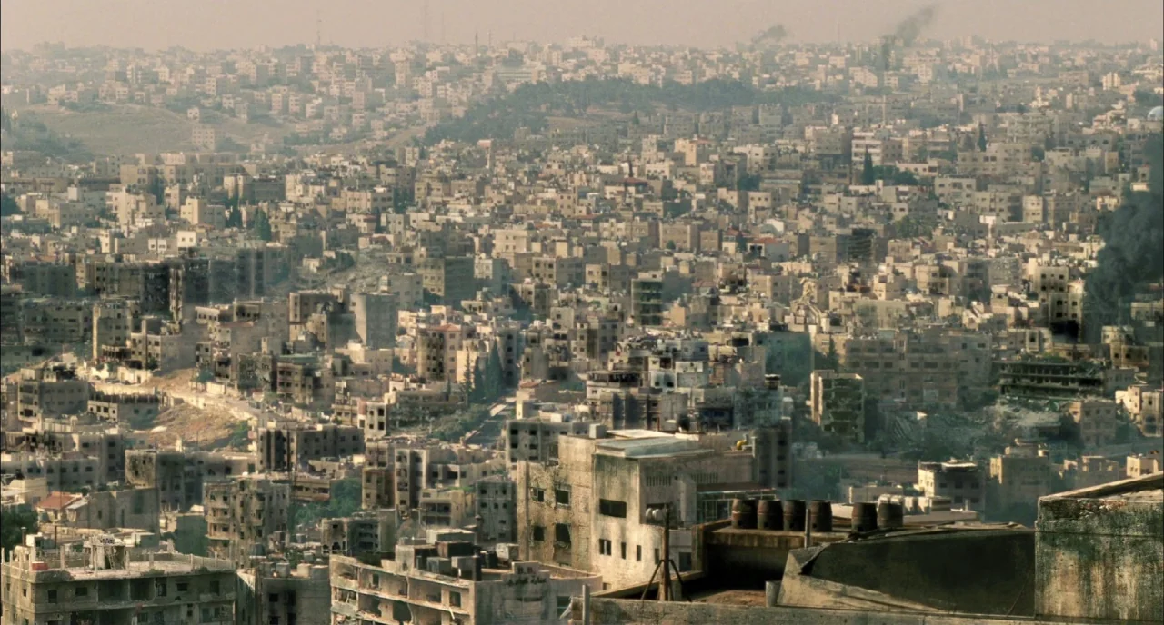 Incendies movie still: establishing — An extensive aerial view looks across a densely packed urban landscape of beige buildings. Thick plumes of…