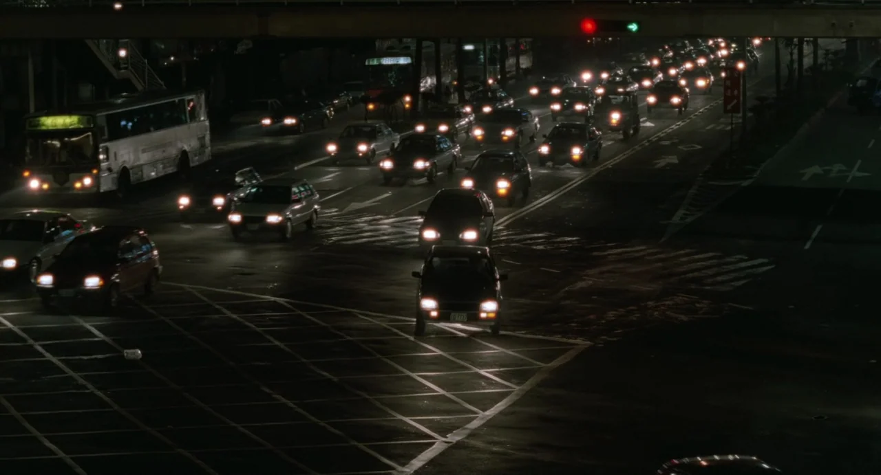 A Confucian Confusion (1994) movie still: establishing — A busy city intersection with multiple lanes of traffic at night. Numerous cars and a bus…