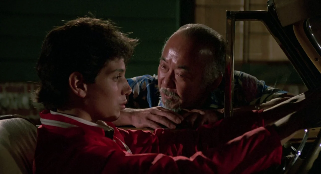 The Karate Kid (1984) movie still: medium — A dark night setting features an older man leaning into the side of a vehicle. He talks to a young man…