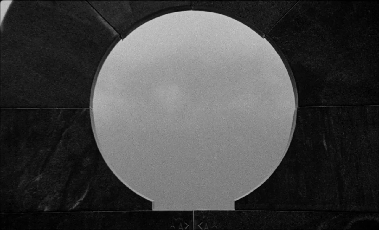 Last and First Men (2020) movie still: closeup — A dark stone slab features a perfect circular cut-out against a bright, hazy sky. The stone has a…