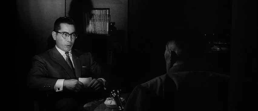The Bad Sleep Well (1960) movie still: medium — An dimly lit office features a man seated at a desk facing another person. The man wears glasses and…