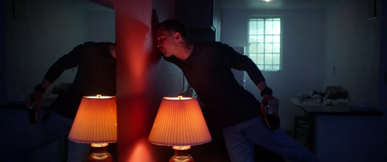 Blindfire (2020) movie still: medium — An interior space features a wall partition illuminated by a single warm desk lamp. A man stands leaning…
