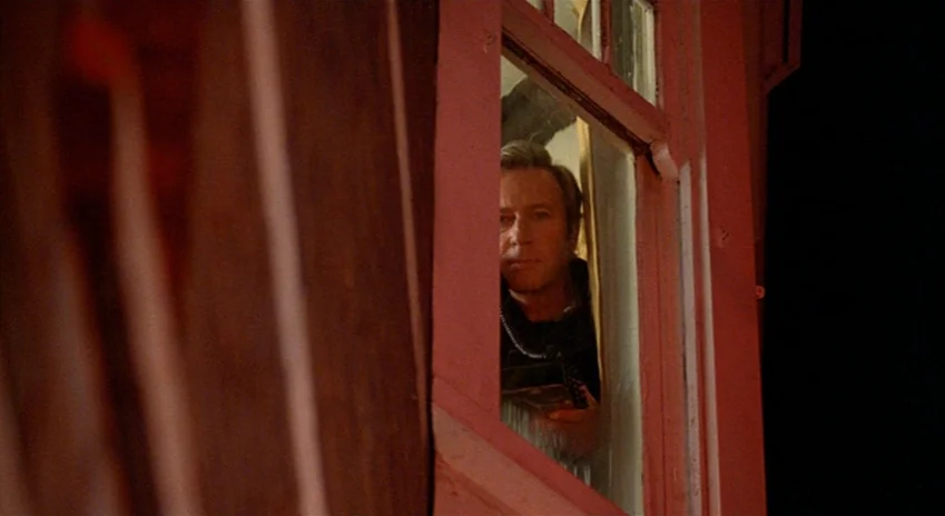 The Silent Partner (1978) movie still: medium — A wooden window frame painted red surrounds a man peering through the glass. The man stands inside…