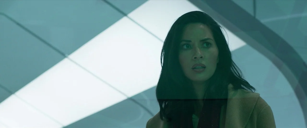 The Predator (2018) movie still: closeup — A woman stands in a modern, brightly lit corridor with curved architectural panels. She looks towards the…
