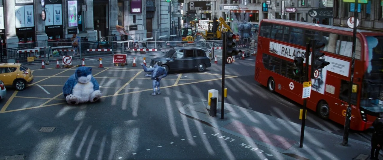Pokémon Detective Pikachu movie still: wide — An urban London intersection features a red double-decker bus and a black taxi cab. Two Pokémon…
