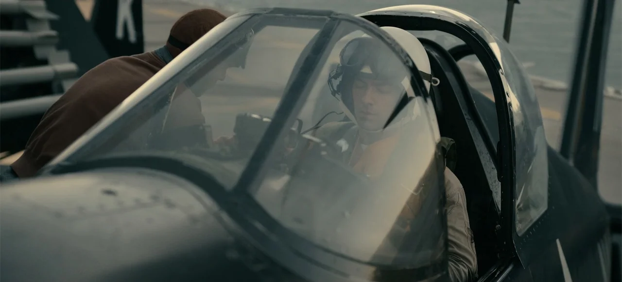 Devotion (2022) movie still: medium — An open cockpit of a vintage fighter plane on an aircraft carrier deck. A pilot in flight gear occupies the…
