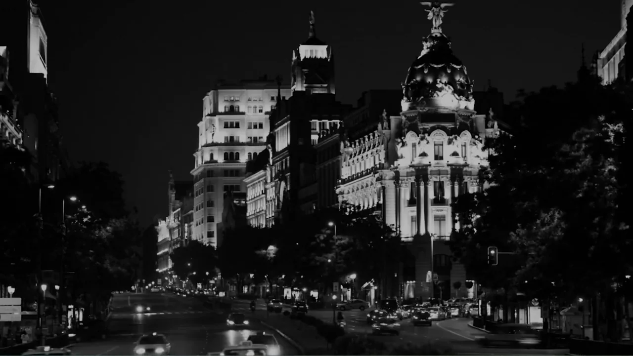 Creditors (2025) movie still: establishing — A night view of a European city intersection with ornate historical architecture illuminated. Cars…