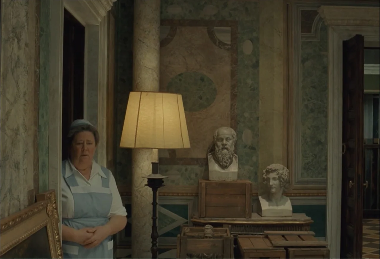 The Phoenician Scheme (2025) movie still: medium — A room with patterned marble walls features stone busts placed atop wooden crates. A woman in a…