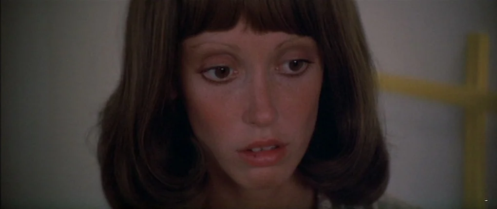 3 Women (1977) movie still: closeup — A woman stands in a room with a plain white wall and a yellow headboard. She stares forward while holding her…