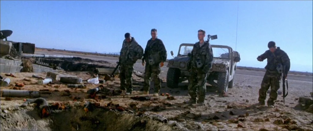 Three Kings (1999) movie still: fullbody — Four soldiers stand in a dry, barren desert landscape near a parked military vehicle. The ground is…