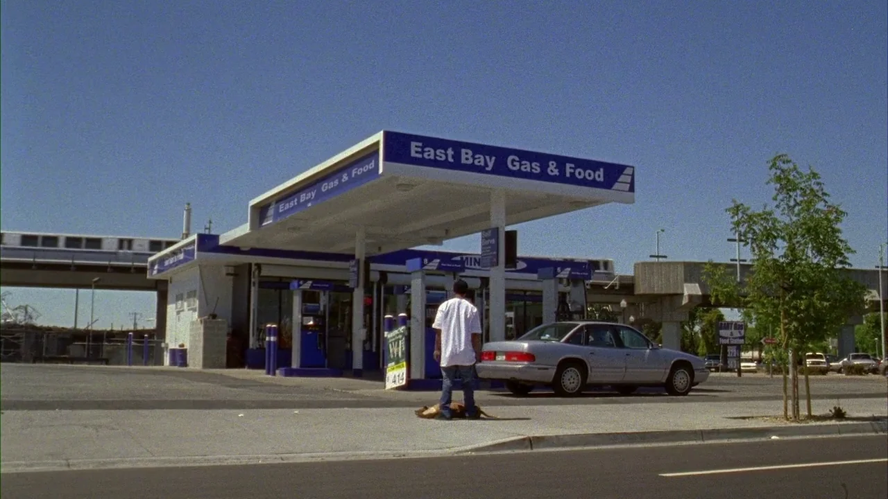 Fruitvale Station (2013) movie still: wide — A roadside gas station featuring a white canopy and a gray sedan parked nearby. An elevated train track…
