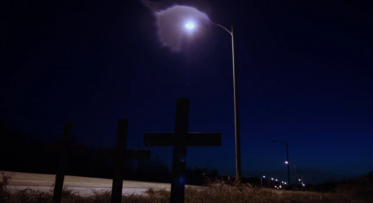 Killer Joe (2011) movie still: wide — A rural roadside at night featuring three wooden crosses set in tall dry grass. A series of bright…