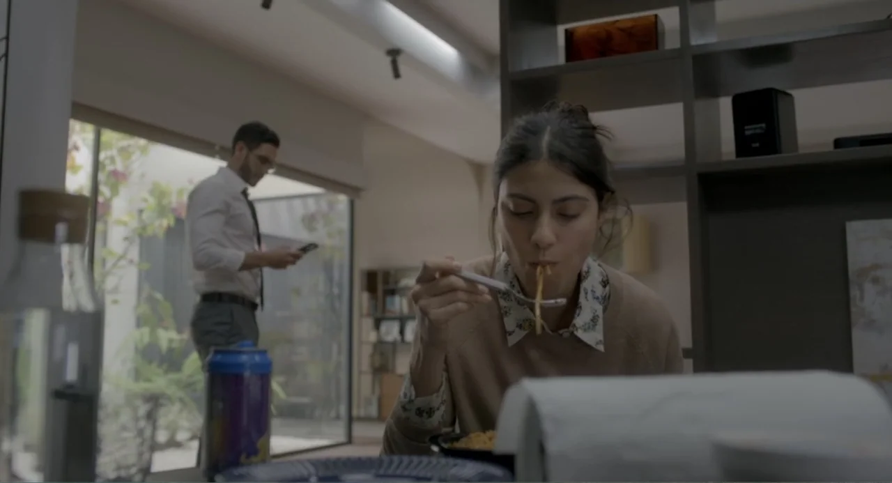 Straight (2024) movie still: medium — A contemporary apartment interior features a woman at a table in the foreground. A man stands in the…