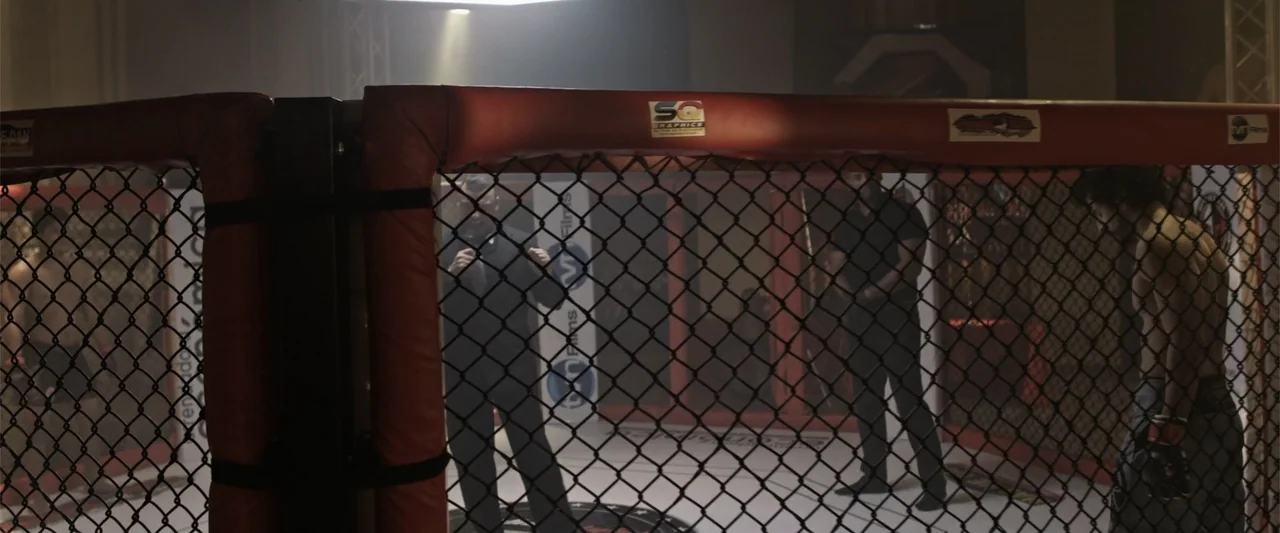 Kimura (2017) movie still: medium — A professional fighting cage interior has red padded corner posts and black chain-link fencing. A referee and a…