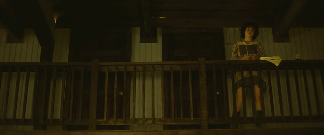 Blaze (2018) movie still: wide — A wooden balcony features a dark railing and two doors set in pale siding. A woman stands on the balcony holding a…