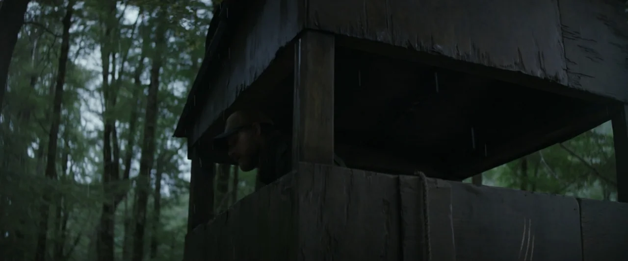Wolf Man (2025) movie still: medium — A dark wooden elevated hunting platform stands amidst a rainy, dense forest of tall trees. A man wearing a cap…