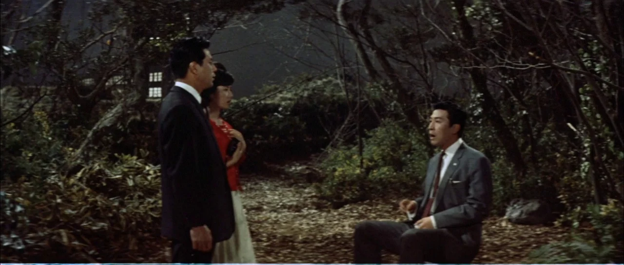 Mothra Vs Godzilla (1964) movie still: fullbody — A clearing in a dense forest at night with fallen leaves covering the ground. Three people gather…