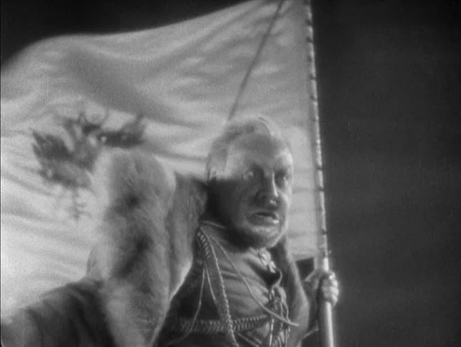 The Last Command (1928) movie still: medium — A dark, atmospheric setting at night with a large flag visible in the background. An older man in…