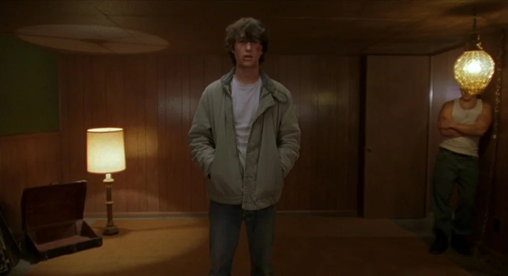 Brick (2005) movie still: medium — A basement with dark wood-paneled walls and a brown carpet floor. Two men stand in the dim room near a vintage…
