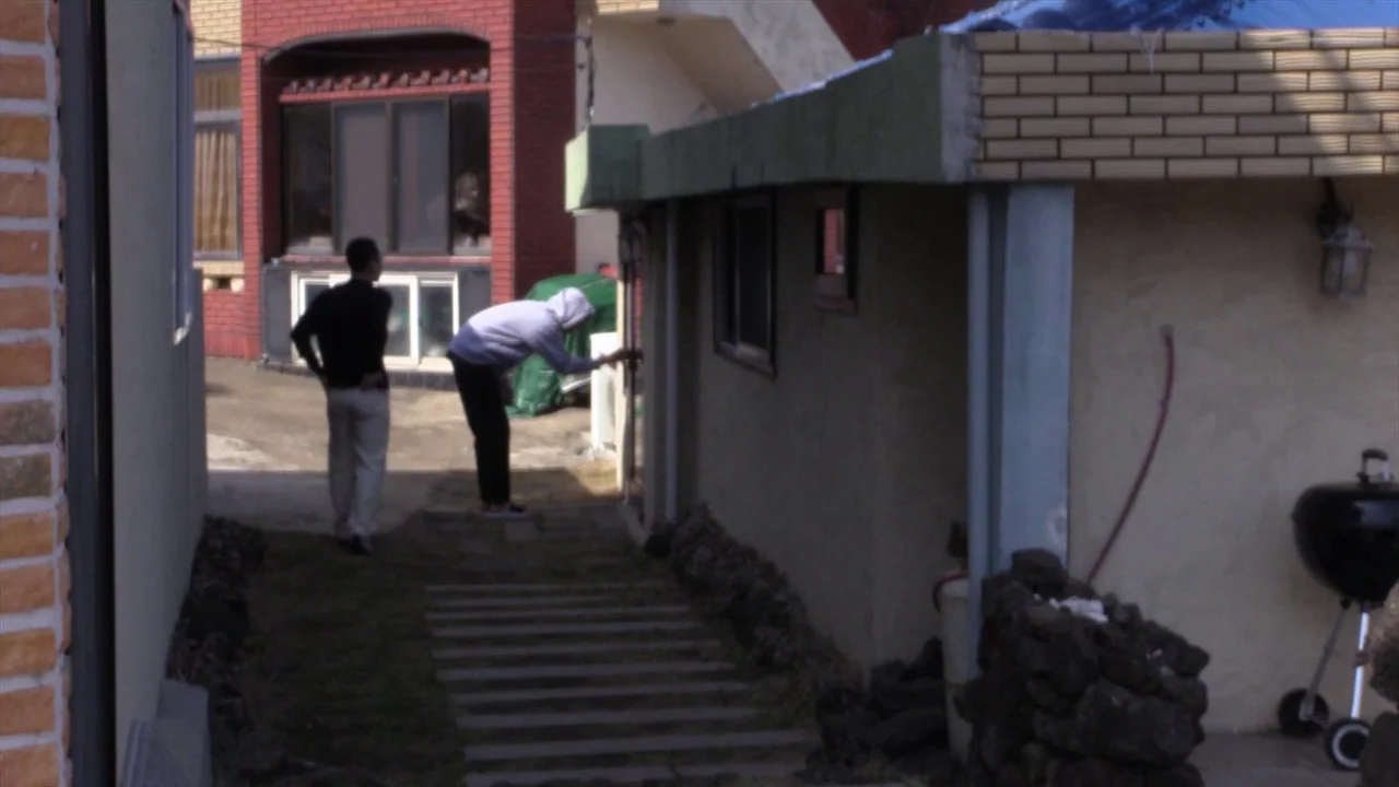 In Water (2023) movie still: wide — A narrow residential courtyard features a paved walkway between brick and stucco buildings. One man stands while…