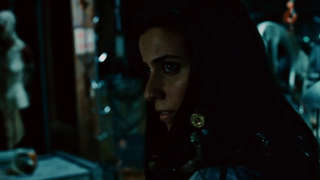 Saw III (2006) movie still: closeup — A dark workshop space features cluttered equipment and deep shadows throughout the environment. A woman with…