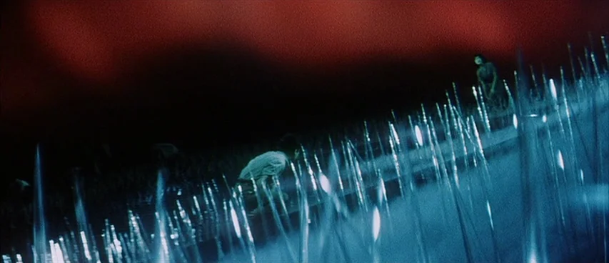Jigoku (1960) movie still: wide — A dark field filled with tall, glowing, translucent spikes under a deep red sky. Two distant figures are visible…