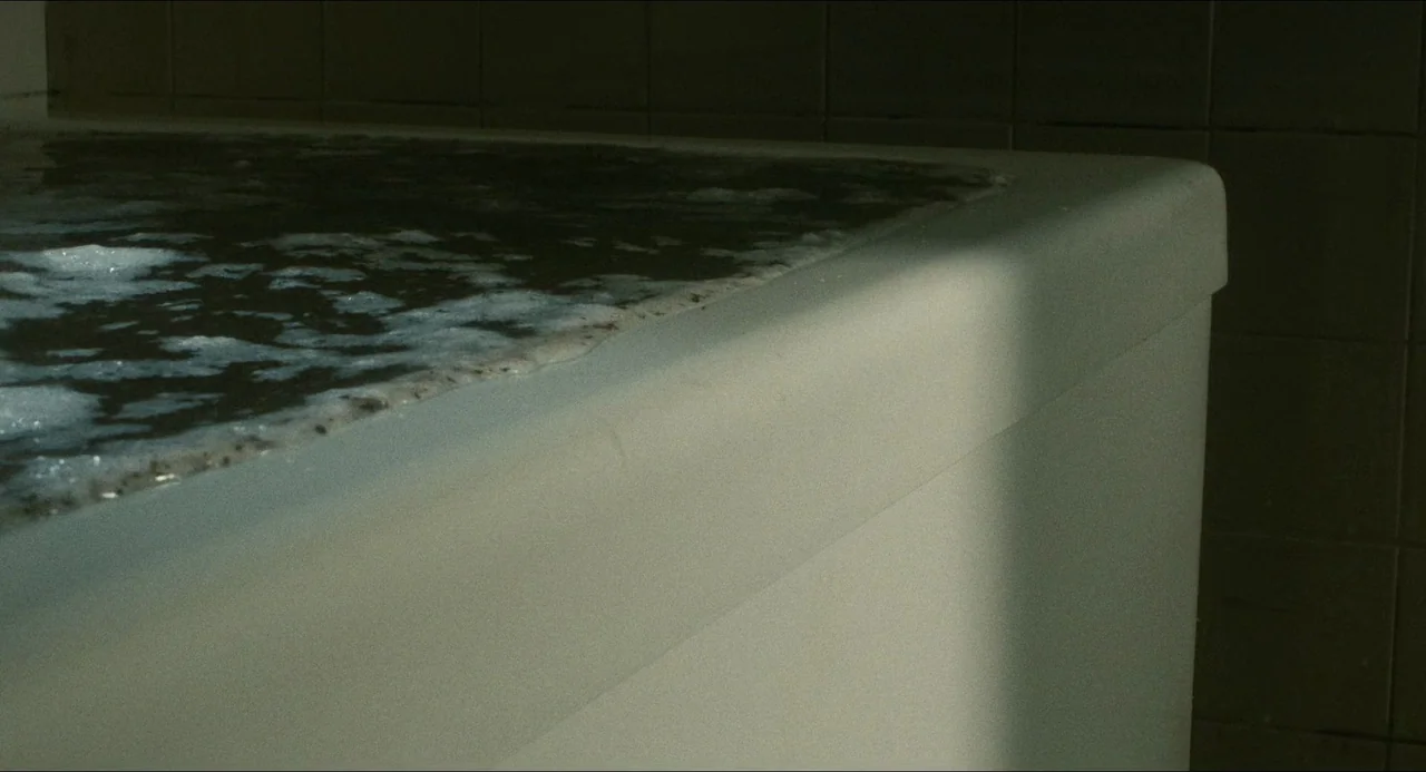Dark Water (2002) movie still: closeup — A dimly lit tiled bathroom features a plain white porcelain bathtub. Dark, murky water with white soap foam…