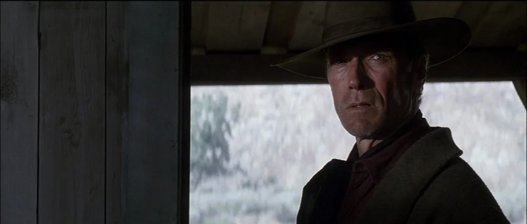 Unforgiven (1992) movie still: medium — A dark, shaded wooden porch looks out onto an overcast and barren natural landscape. An older man wearing a…