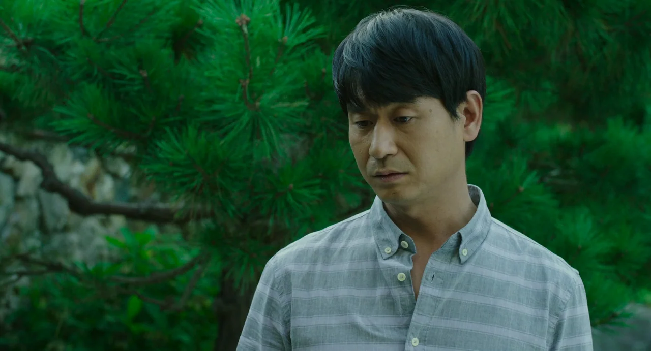 The Mimic (2017) movie still: medium — An outdoor area is filled with dense green pine tree branches and foliage. A middle-aged man in a striped…