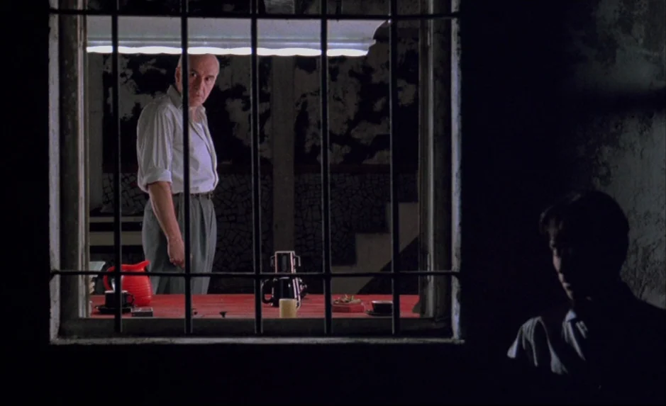 Mauvais Sang (Bad Blood) (The Night Is Young) (1986) movie still: medium — A dimly lit exterior view looks through a barred window into a brightly…