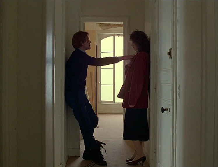 First Name: Carmen (1983) movie still: fullbody — A narrow residential hallway with white walls and a door leading to a brighter room. A man faces a…