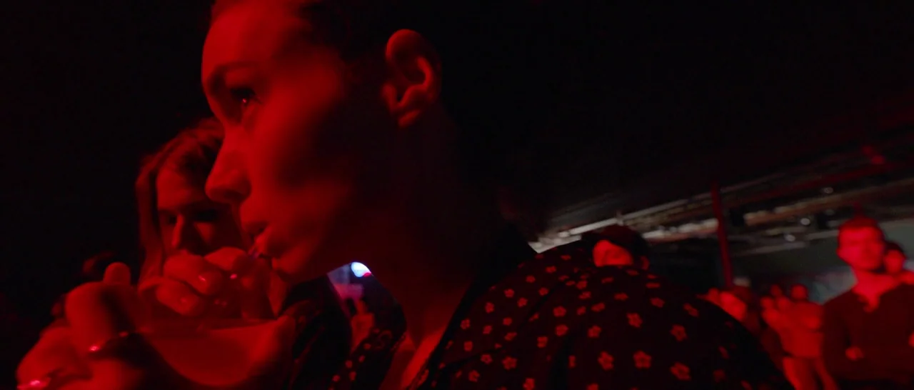 Song to Song (2017) movie still: closeup — A dimly lit nightclub interior filled with deep red light. A young woman drinks from a glass while…