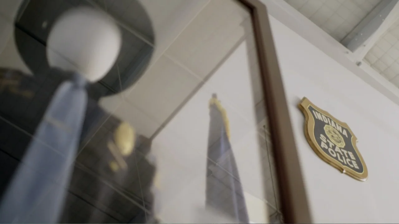 Murders at the Burger Joint (2022) movie still: medium — An Indiana State Police emblem is mounted on a white wall near a glass case. The glass case…