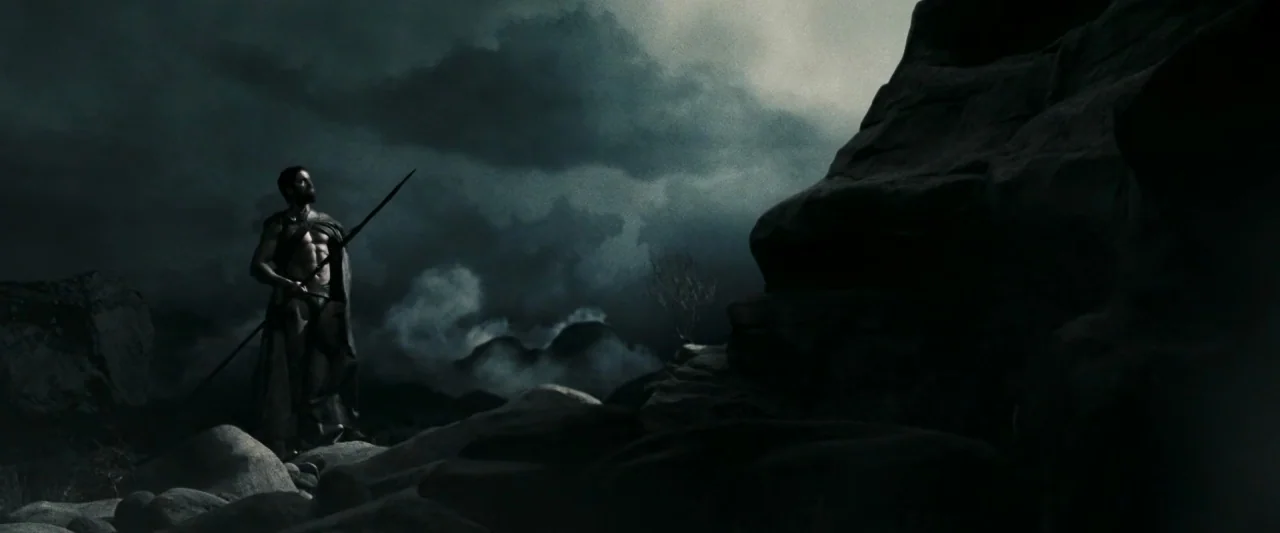 300 (2006) movie still: wide — A rugged rocky terrain under a dark, storm-filled sky with thick clouds. A lone warrior stands holding a spear while…