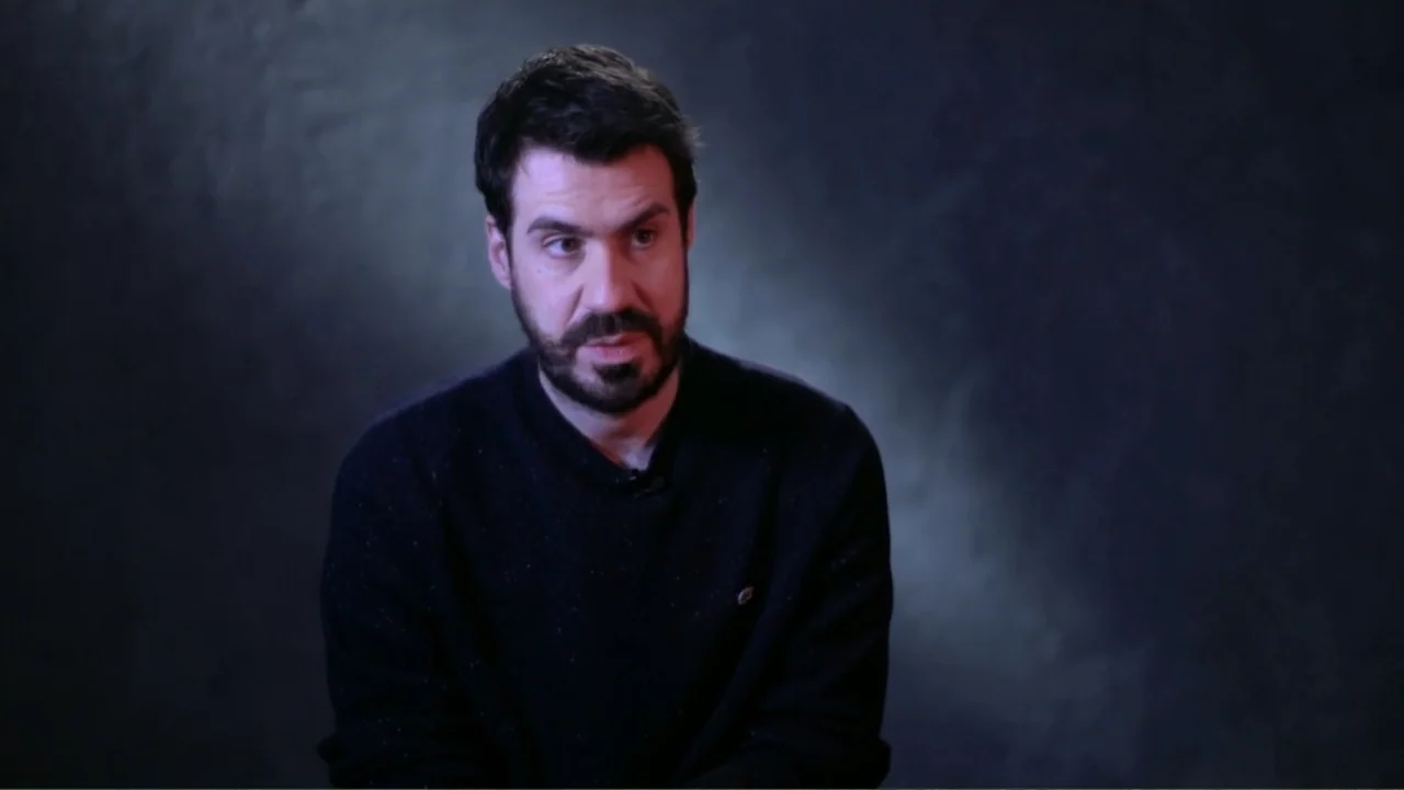 Reign of the Superwomen (2021) movie still: medium — A man sits in front of a dark, mottled studio background. He faces slightly right while talking…