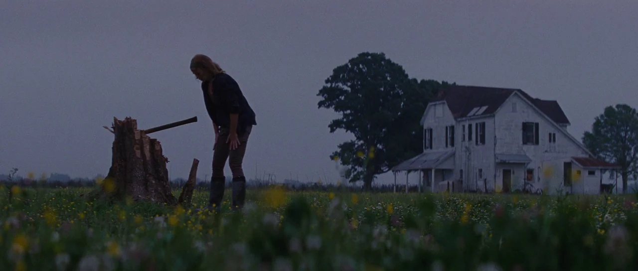 Looper (2012) movie still: wide — A woman stands in a vast, grassy field under a grey, overcast sky. She leans toward a wooden tree stump with an…