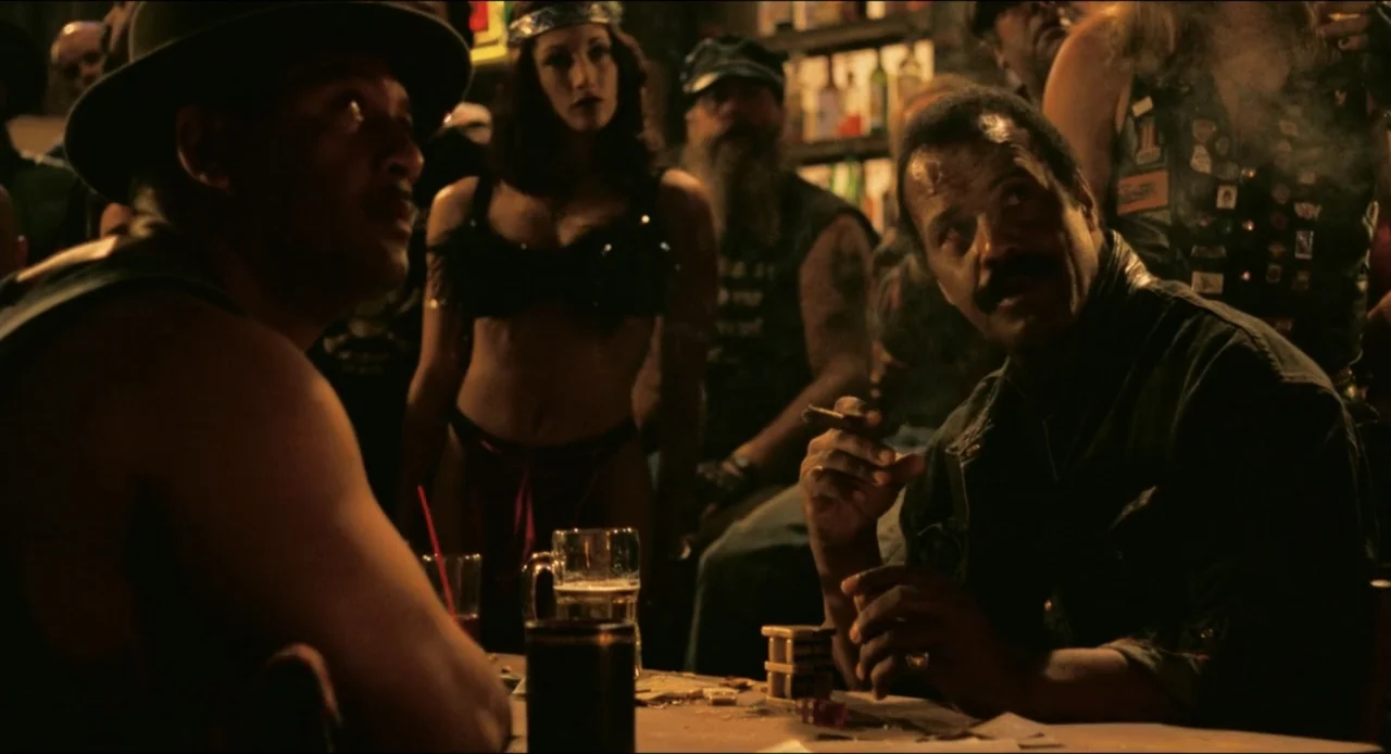 From Dusk Till Dawn (1996) movie still: medium — A dark, wood-paneled bar is filled with people in leather attire and smoke. Two men sit at a…