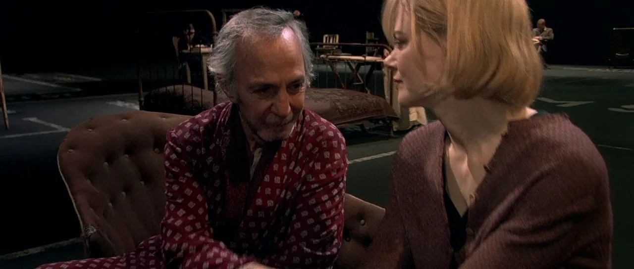Dogville (2003) movie still: medium — A stark minimalist set features isolated furniture pieces against a pitch-black background. A man in a…