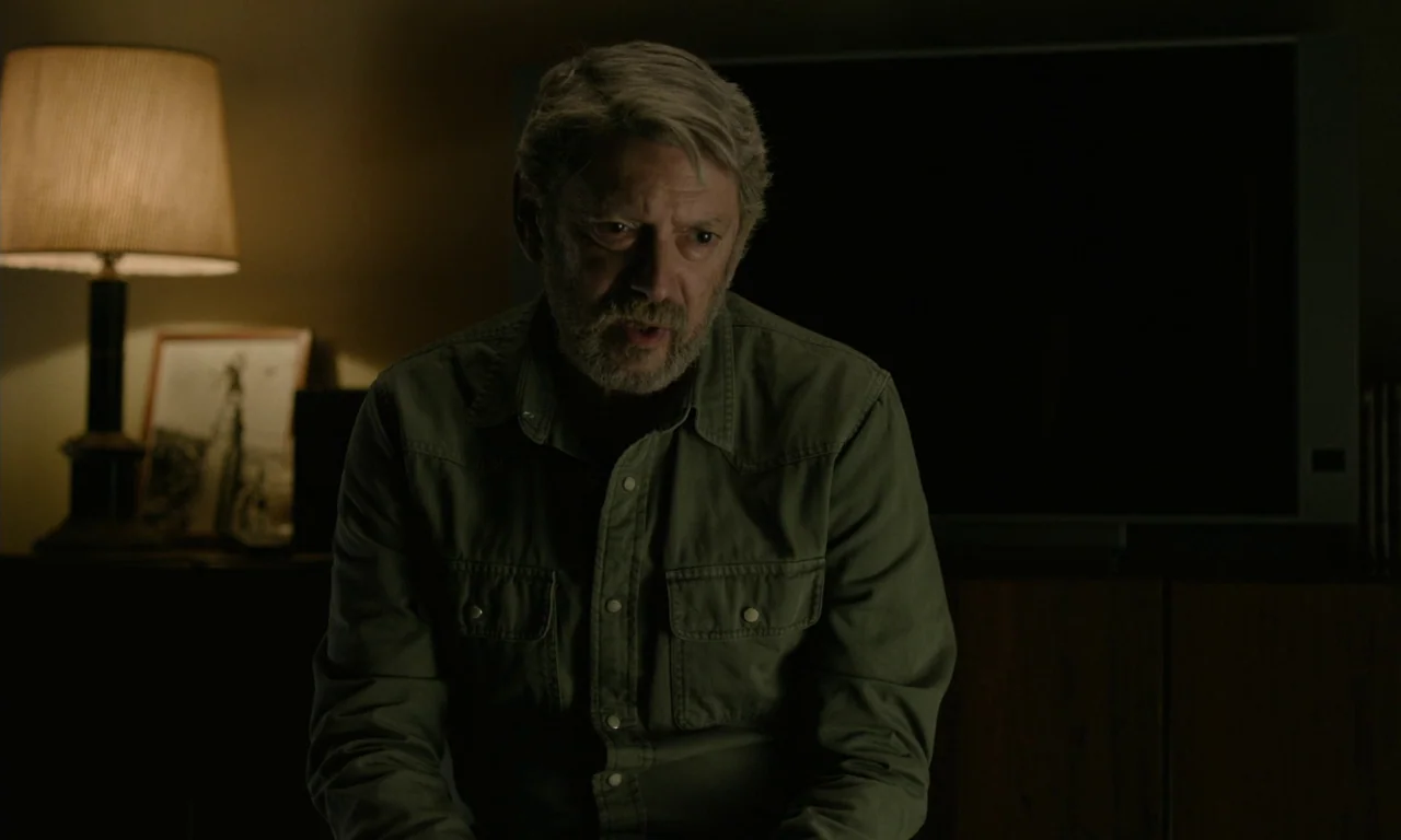 Close Your Eyes (2023) movie still: medium — A dimly lit room at night contains a dark television and a bedside lamp. An older man with a gray beard…