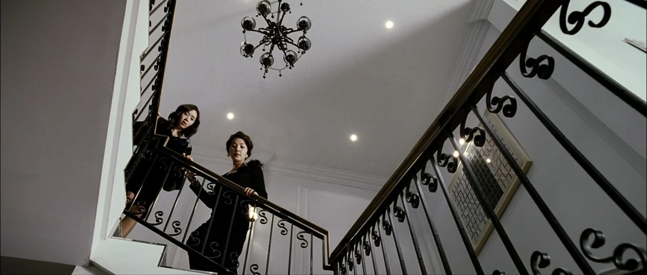 The Housemaid (2010) (2010) movie still: medium — Two women descend a white modern staircase with ornate black iron railings. A decorative…