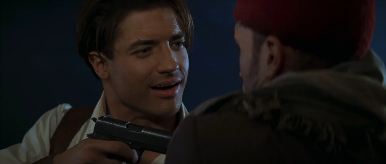 The Mummy (1999) movie still: medium — A dark indoor space provides the backdrop for a confrontation between two men. One man aims a firearm at the…