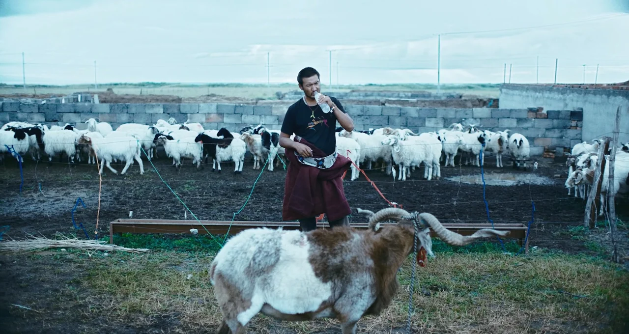 Balloon (2019) movie still: medium — An outdoor sheep pen with a grey block wall under a cloudy sky. A man stands in the middle of the pen drinking…