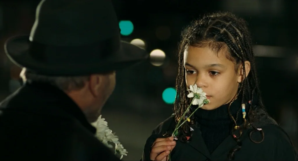 This Night (2008) movie still: medium — A dark night street setting features a girl and a man facing each other. The young girl holds white flowers…