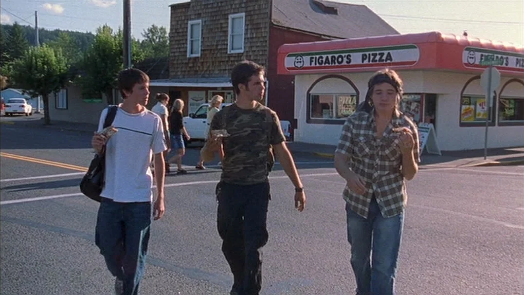 Mean Creek (2004) movie still: fullbody — An intersection in a small town features a pizza restaurant and wooden buildings. Three teenage boys walk…