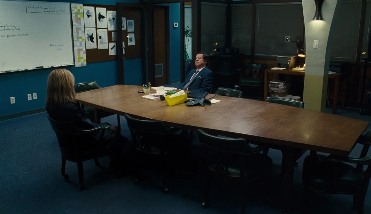 Our Brand is Crisis (2015) movie still: wide — A dimly lit corporate boardroom with blue walls and a long wooden conference table. A man in a suit…