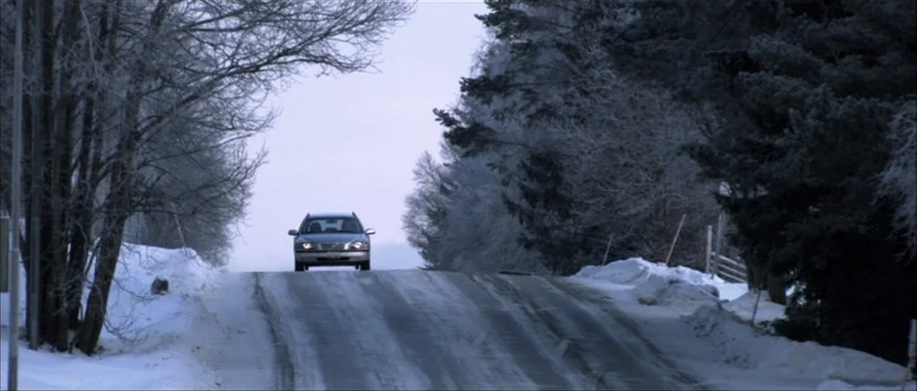 Genova (2008) movie still: wide — A remote road stretches through a forest covered in heavy winter frost and snow. A single silver station wagon…