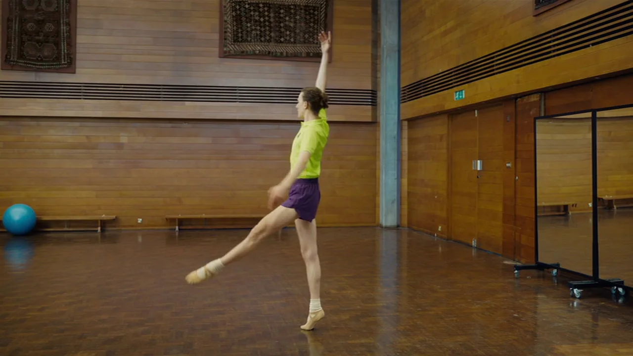 Club Zero (2023) movie still: fullbody — A spacious room features polished wooden floors and walls lined with horizontal wooden slats. A male dancer…
