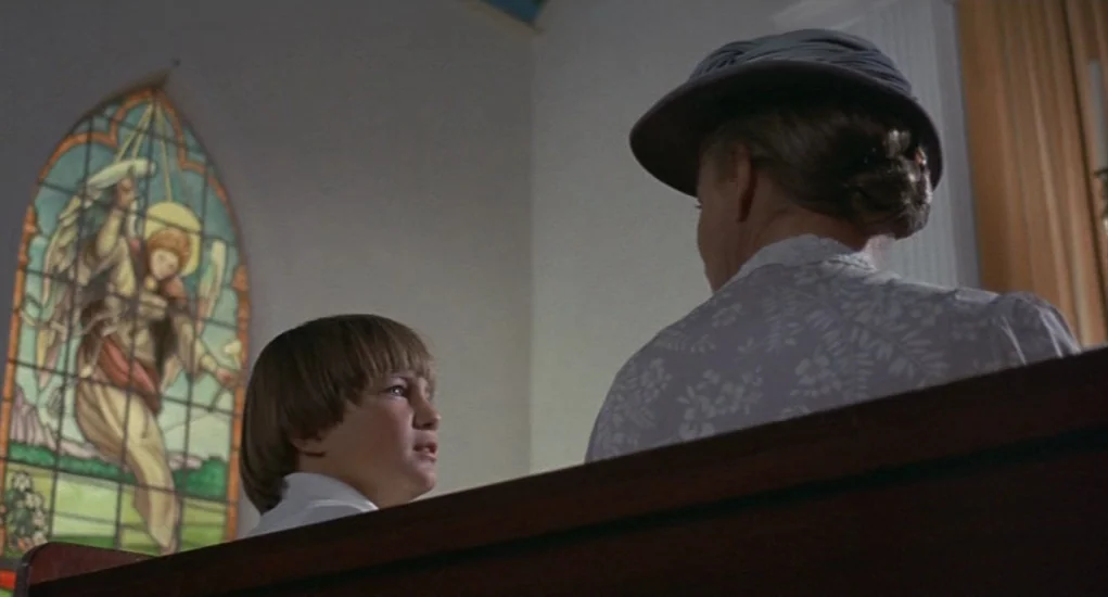 The Other (1972) movie still: medium — A church interior with a wooden pew and a prominent stained glass window. A young boy looks toward an elderly…